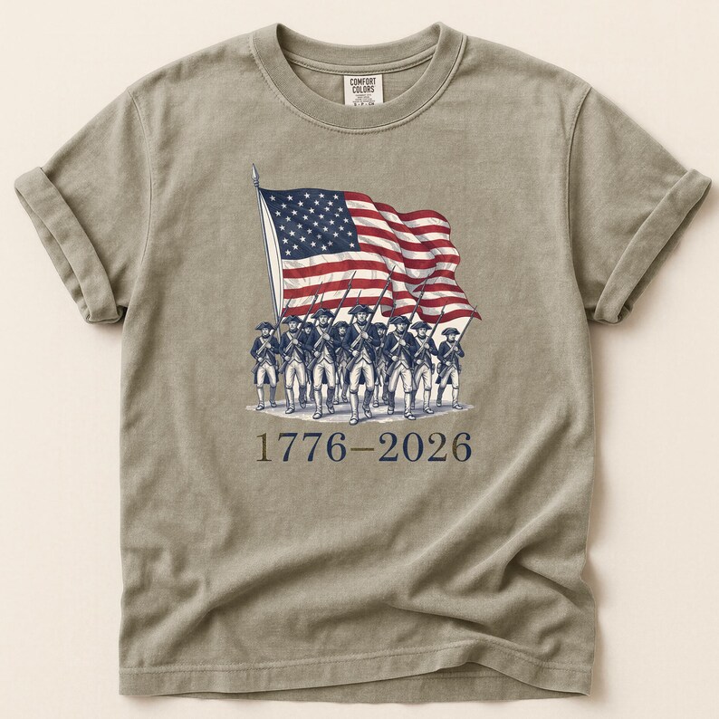May include: A tan t-shirt featuring a vintage illustration of the American flag waving above a line of soldiers. The text "1776-2026" is printed below the image. The shirt has rolled-up sleeves and a crew neck.