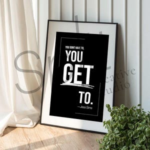 May include: A framed black and white print with the text "YOU DON'T HAVE TO, YOU GET TO." The print is in a black frame and is leaning against a white wall. A white curtain and a small green plant are also visible.