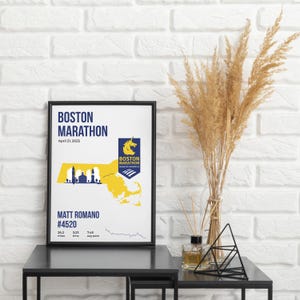 May include: Framed Boston Marathon poster with a black frame. The poster features the words "BOSTON MARATHON" and "April 21, 2025". It includes a yellow and blue map of Massachusetts, the Boston Marathon logo, and runner details.