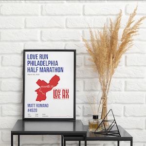May include: A framed poster with the text "LOVE RUN PHILADELPHIA HALF MARATHON" in blue. The poster includes a red map outline and the name "MATT ROMANO #4520". The poster is on a black table with decorative items.