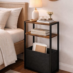 May include: A three-tiered side table with a black metal frame and wooden shelves. The top shelf holds a white lamp, decorative items, and books. The bottom shelf has a dark gray fabric bin for storage. A neutral rug is in the foreground.
