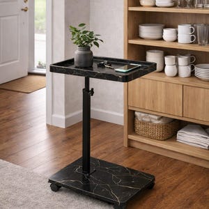 May include: A black marble-patterned adjustable side table with a rectangular top and base, supported by a black metal pole. The table has wheels and holds a small plant, keys, and a smartphone. The table is next to a wooden cabinet.
