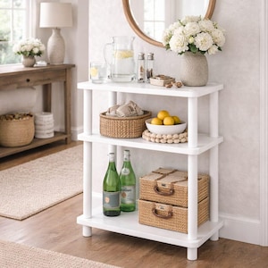 May include: A white three-tiered shelf unit displays decorative items. A round mirror hangs above, and a vase of white flowers sits on the top shelf. Other items include a woven basket, lemons, and two green bottles. Two woven storage boxes are on the bottom shelf.