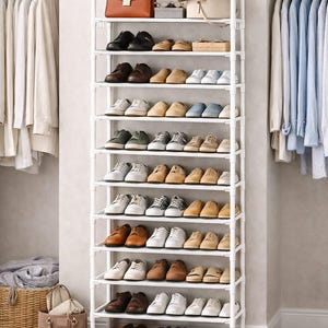 May include: A white metal shoe rack filled with various shoes, a brown purse, and a tan handbag. The shoes are in different styles and colors, including black, brown, white, and tan. The rack is in a closet with hanging clothes.