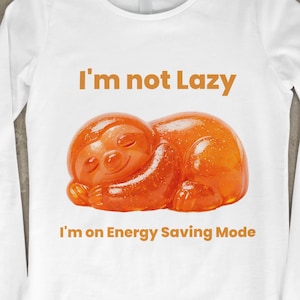 May include: White long-sleeve shirt with the text "I'm not Lazy" and "I'm on Energy Saving Mode". The shirt features an orange sloth graphic. The shirt is made of a soft, comfortable material.