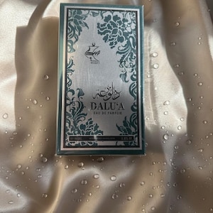 Puede incluir: Caja de perfume rectangular con un diseño plateado y verde azulado. La caja presenta el texto "DALU'A Eau de Parfum" y la marca "MUSKY". La caja está sobre una superficie satinada.