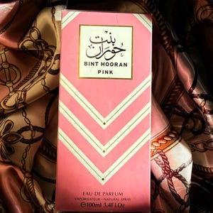 Puede incluir: Caja de perfume rosa con detalles blancos y dorados. La caja muestra el texto "BINT HOORAN PINK" en inglés y árabe. Contiene 100 ml de Eau de Parfum.