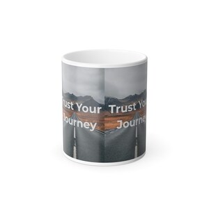 May include: White ceramic coffee mug featuring a scenic road design with the text "Trust Your Journey" in white. The design includes a road leading towards mountains under a cloudy sky.