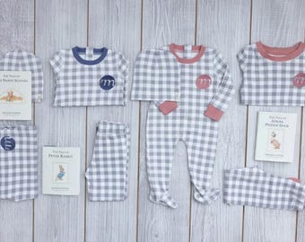 May include: A collection of gray and white gingham baby clothing including long-sleeve shirts, pants, and hats with pink or blue accents and circular monogram patches, accompanied by three small Beatrix Potter storybooks.