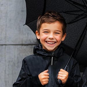 May include: A smiling child in a black hooded jacket holding a black umbrella with a leopard print design. The umbrella is open. The child is looking at the camera.