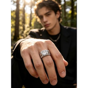 May include: A silver-toned ring featuring a prominent, clear, round-cut center stone set within a square-shaped bezel. The band displays intricate, textured detailing along the sides, providing a distinct contrast to the polished metal finish.