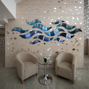 May include: A modern interior with two beige armchairs, a small glass table, and a decorative wall art piece. The wall art features blue and teal wave-like shapes against a light wood-toned background with irregular cutouts.