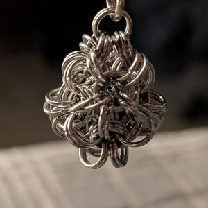 Könnte beinhalten: Ein silberfarbener, kugelförmiger Anhänger aus ineinandergreifenden Metallringen. Der Anhänger ist an einem kleinen Metallverschluss und einem Ring befestigt. Das Design ist aufwendig und scheint handgefertigt zu sein.