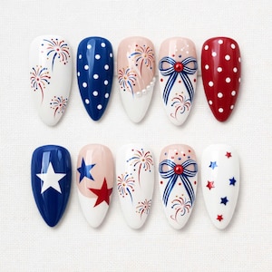 May include: A set of ten artificial nails with patriotic designs. The nails feature white, red, and blue colors with patterns including fireworks, polka dots, stars, and bows. The nails are almond-shaped and arranged on a white surface.