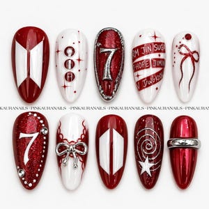 May include: A collection of ten artificial nails in red and white. The designs include geometric patterns, text, and embellishments. Some nails display the number "7" and the names "RM JIN SUGA T-HOPE JIMIN VJWGKOOK". The nails are designed for cosmetic use.