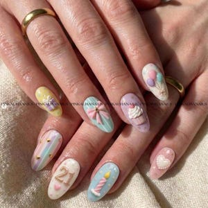 May include: A set of long, oval-shaped nails with various 3D designs. Designs include a bow, cupcake, balloons, a candle, and the number "21". Colors include pastel shades of pink, blue, yellow, and purple. Gold accents are also present.