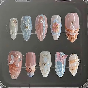 May include: A set of ten artificial nails in a clear plastic case. The nails feature various designs, including iridescent pink and blue hues, floral accents, and seashell-inspired details. Some nails have 3D embellishments.