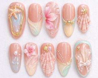 May include: A set of ten almond-shaped artificial nails with a beach theme. Each nail features a different design, including a starfish, flowers, seashells, and pearls, in shades of pink, gold, and blue. The nails are decorated with 3D elements.