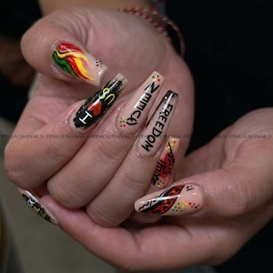 Può includere: Una manicure con unghie acriliche lunghe e quadrate, decorate con vari disegni. Un'unghia presenta un cuore rosso e il testo "I US". Altre unghie mostrano le parole "NMMCA" e "FREEDOM". Le unghie sono dipinte con accenti rossi, gialli, verdi e neri.