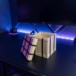 May include: A small, open wooden crate with a hinged lid, secured by black bands. The crate is made of light-colored wood and sits on a black surface. The lid is partially open, revealing the interior of the crate. The background includes a computer monitor and desk.
