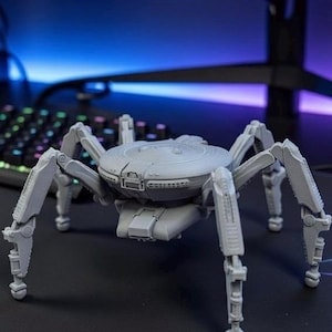 Może przedstawiać: Szary, mechaniczny model pająka z przegubowymi nogami i szczegółowym korpusem. Pająk jest umieszczony na czarnej powierzchni, z klawiaturą i monitorem komputera w tle. Model ma futurystyczny, robotyczny design.