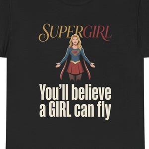 Puede incluir: Camiseta negra con el logo de Supergirl y un gráfico de Supergirl. El texto "You'll believe a GIRL can fly" está impreso debajo. La camiseta está hecha de un material suave.
