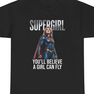 Puede incluir: Camiseta negra con la palabra "SUPERGIRL" en letras plateadas sobre una ilustración de Supergirl con su disfraz. El texto "YOU'LL BELIEVE A GIRL CAN FLY" está debajo. La camiseta está hecha de un material suave.