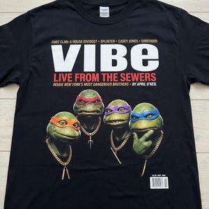May include: Black t-shirt featuring the Teenage Mutant Ninja Turtles. The shirt has the word "VIBE" in large white letters, with the text "LIVE FROM THE SEWERS" below. The image includes the turtles with gold chains.