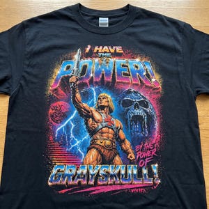 May include: Black t-shirt featuring a colorful graphic of He-Man holding a sword, with the text "I HAVE THE POWER!" and "BY THE POWER OF GRAYSKULL!". The design includes a skull, lightning, and planets.