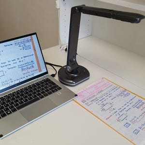 Puede incluir: Un portátil que muestra ecuaciones matemáticas manuscritas está sobre un escritorio blanco, junto a una hoja de papel con ecuaciones similares. Una lámpara de escritorio negra y ajustable está colocada encima. La escena sugiere un entorno de estudio o trabajo.