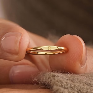 May include: A thin gold ring featuring a small, flat, oval-shaped centrepiece engraved with the letter R. The band is smooth and polished, showcasing a minimalist design with a reflective metallic finish that catches the light.