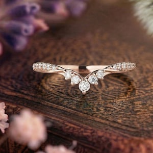 Könnte beinhalten: Ein V-förmiger Ring aus Roségold, besetzt mit einer Reihe kleiner, runder, klarer Steine entlang der Ringschiene. Das Design umfasst einen zentralen Stein an der Spitze des V-förmigen Bogens sowie weitere Steine entlang der beiden Seiten.