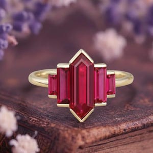 Könnte beinhalten: Ein goldener Ring mit einem länglichen, roten Edelstein in der Mitte, flankiert von kleineren, rechteckigen roten Steinen. Der Ring hat ein geometrisches Design, vor einem verschwommenen Hintergrund aus Blumen und einer Holzoberfläche. Das Design des Rings ist vom Art Déco inspiriert.