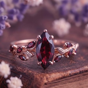 Könnte beinhalten: Roségoldener Ring mit einem großen, marquiseförmigen, tiefroten Edelstein als Mittelpunkt. Das Band ist mit kleineren roten Edelsteinen und blattartigen Designs verziert. Der Ring ist vor einem verschwommenen Hintergrund aus Blumen platziert.