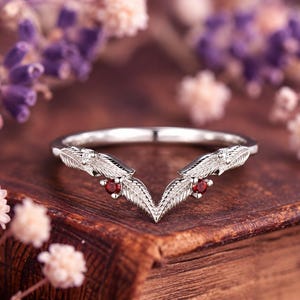 May include: A silver-toned V-shaped ring featuring a delicate leaf-patterned band. The design includes two small, round, deep red gemstones set symmetrically near the center point, accented by tiny clear stones embedded within the metal leaves.