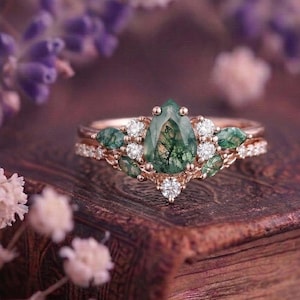 Könnte beinhalten: Ein Ring aus Roségold mit einem zentralen, birnenförmigen Moosachat mit dunkelgrünen Einschlüssen, umgeben von kleineren, marquise-geschliffenen Moosachatsteinen und runden, weißen Brillanten, die in ein zartes Metallband gefasst sind.