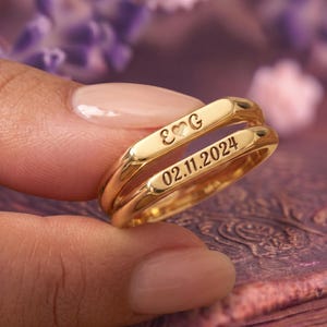 Könnte beinhalten: Zwei goldfarbene Ringe mit flachen, polierten Oberflächen. Der obere Ring ist mit den Buchstaben E und G graviert, die durch ein Herzsymbol getrennt sind. Der untere Ring trägt die Gravur des Datums 02.11.2024 in einer klaren, dunklen Schrift.
