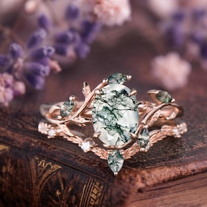 Könnte beinhalten: Ein roségoldenes Ring-Set mit einem oval geschliffenen Moosachat-Mittelstein mit grünen Einschlüssen. Der Ring ist mit Blatt- und Rankendetails und kleinen, klaren Edelsteinen verziert. Das Band hat ein zartes, von der Natur inspiriertes Design.