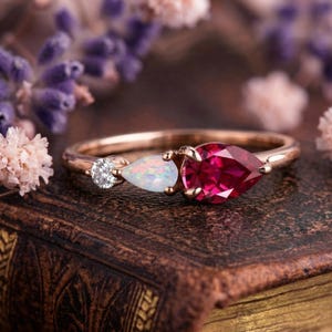 Könnte beinhalten: Ein roségoldener Ring mit drei Edelsteinen. Links befindet sich ein kleiner, runder, klarer Edelstein, gefolgt von einem birnenförmigen Opal und rechts einem größeren, birnenförmigen, tiefroten Edelstein. Der Ring ist vor einem verschwommenen Hintergrund aus lila und rosa Blumen platziert.