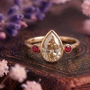Könnte beinhalten: Ein goldener Ring mit einem birnenförmigen, hellfarbigen Edelstein als Mittelpunkt, flankiert von zwei kleineren, runden roten Edelsteinen. Der Ring ist vor einem verschwommenen Hintergrund aus lila und rosa Blumen platziert.