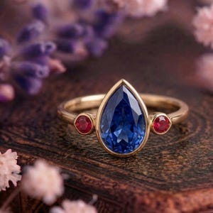 Könnte beinhalten: Ein goldener Ring mit einem großen, tropfenförmigen, tiefblauen Edelstein im Zentrum, flankiert von zwei kleineren, runden, roten Edelsteinen. Der Ring hat ein schlichtes, poliertes Goldband. Der Hintergrund ist verschwommen, mit Andeutungen von lila und rosa Blüten.