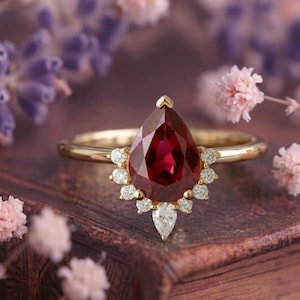 Könnte beinhalten: Ein goldener Ring mit einem großen, birnenförmigen, tiefroten Edelstein. Der Edelstein ist von kleineren, klaren Akzentsteinen umgeben. Der Ring hat ein schlichtes, poliertes Goldband. Der Ring ist vor einem verschwommenen floralen Hintergrund platziert.