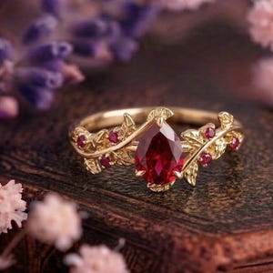 Könnte beinhalten: Ein goldener Ring mit einem birnenförmigen, tiefroten Edelstein als Mittelpunkt. Der Ring hat ein florales Design mit kleinen roten Edelsteinen und Blattakzenten. Der Ring ist vor einem verschwommenen Hintergrund aus Blumen und einer Holzoberfläche platziert.
