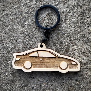 Könnte beinhalten: Ein holzfarbener Schlüsselanhänger in Form eines Autos mit einem schwarzen Metallring und einer Kette. Das Autodesign ist lasergeschnitten und zeigt die Kontur eines Coupés mit Rädern und Fenstern. Der Schlüsselanhänger ist aus hellem Holz gefertigt und liegt auf einer strukturierten grauen Oberfläche.