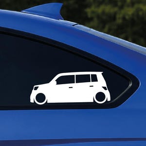 Peut inclure: Un autocollant en vinyle blanc représentant la silhouette latérale d'une voiture à hayon carrée avec une suspension abaissée et de grandes roues, appliqué sur la vitre latérale arrière teintée d'un véhicule bleu vif sur fond forestier flou.