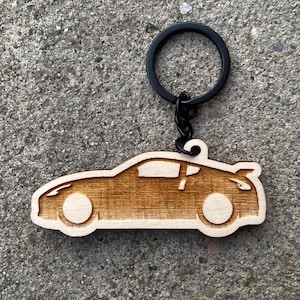 Può includere: Un portachiavi in legno a forma di auto con un portachiavi nero. Il portachiavi è marrone chiaro e presenta un contorno di auto, comprese le ruote e uno spoiler. Il portachiavi è in legno ed è un accessorio per auto.