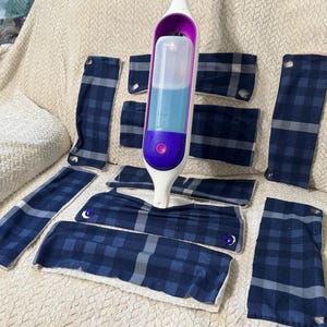 Puede incluir: Una herramienta de limpieza blanca y morada con un recipiente transparente lleno de líquido azul, rodeada de varias almohadillas de limpieza a cuadros azul marino y gris. Las almohadillas son de varios tamaños y formas.