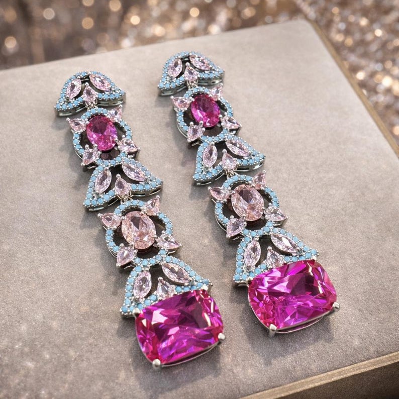Puede incluir: Un par de pendientes largos colgantes con un engaste plateado. Los pendientes presentan una combinaci&oacute;n de piedras preciosas rosas y celeste claro. El dise&ntilde;o incluye piedras ovaladas y rectangulares, creando un accesorio glamuroso.