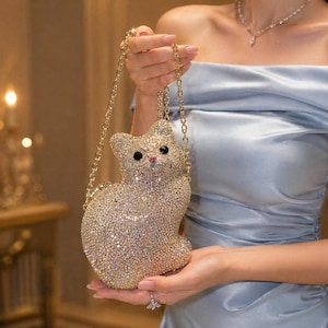 Puede incluir: Un bolso dorado con forma de gato cubierto de brillantes pedrería. El bolso tiene una correa de cadena dorada y ojos negros. El gato está sentado erguido, con la cola enroscada. El fondo está borroso, con una persona sosteniendo el bolso.