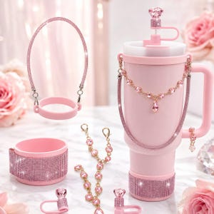 Peut inclure: Un gobelet rose clair avec une poignée, orné d'une base incrustée de strass et d'une chaîne décorative. L'ensemble comprend une protection en silicone rose assortie, une sangle recouverte de strass, une breloque et des accessoires roses.
